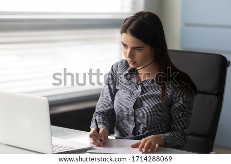 Attractive businesswoman talking using headphones signing up document looking at laptop screen. Serious woman operator working in call center and support people. Manager communicating with client.