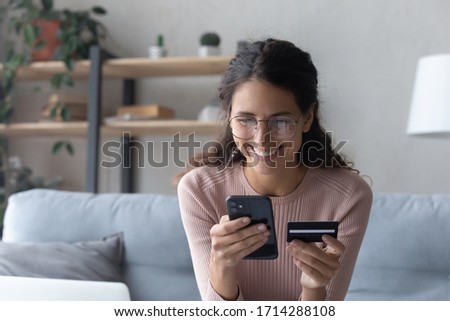 Smiling happy young woman client in eyeglasses enjoying shopping in mobile application, entering information from banking credit card, confirming purchase of delivery food or taxi, online order. Royalty-Free Stock Photo #1714288108