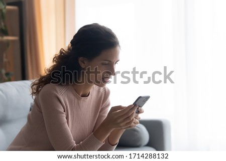 Side view pretty young woman sitting on comfortable couch, looking at mobile phone screen, involved in pleasant conversation in social networks or reading interesting article information on website.
