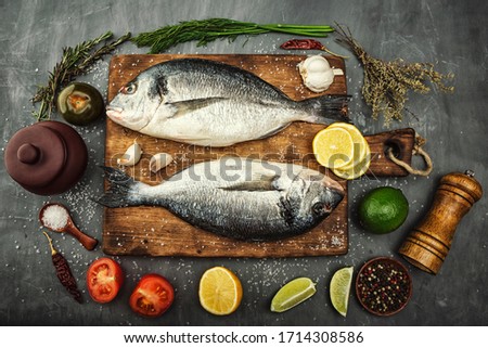 Two Dorado fish lie on a wooden chopping Board on beautiful gray concrete. Next are: lime, lemon, pepper, tomato, garlic, spices, salt and herbs. Royalty-Free Stock Photo #1714308586