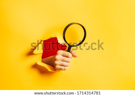 A human hand holds a magnifying glass through a torn hole in a paper yellow wall. Search information concept. Side view for text.