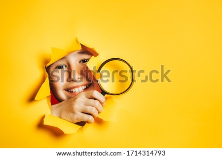 Little smiling caucasian boy looks through torn hole in paper yellow wall, holding magnifying glass in his hand. Search, discovery, research concept. Sidebar for text information, advertising, event. Royalty-Free Stock Photo #1714314793