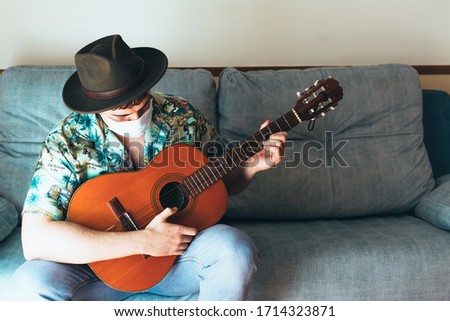Bohemian musician with mask on his face and hat playing the Spanish guitar from the sofa. Dressed in a Hawaiian shirt and jeans. Concept music from home during confinement. Coronavirus pandemic.