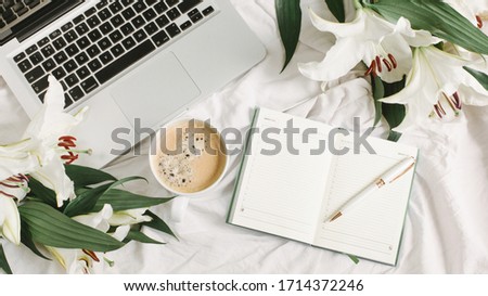 Morning bed with notebook, laptop, coffee cup and flowers on the white bed sheet. Good morning and weekend concept in flat lay style