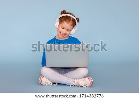 Full body positive little girl in headphones smiling and watching cartoon on modern laptop while sitting crossed legged on blue background
