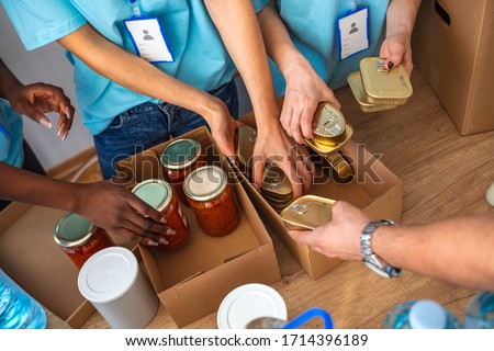 Cardboard boxes being filled with food donations. Helpful team of social workers. Young people volunteering to sort donations for charity food drive. Unrecognizable people volunteering in food bank Royalty-Free Stock Photo #1714396189