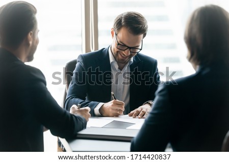 Smiling confident businessman wearing glasses signing contract at group negotiations, business partners making successful investment deal, agreement, salesman putting signature on legal documents Royalty-Free Stock Photo #1714420837