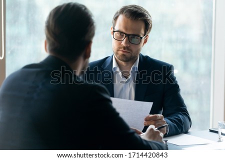 Serious attentive HR manager wearing glasses looking at job applicant, holding cv, listening to candidate on interview, strict employer, recruiter making decision about hiring, difficult negotiations Royalty-Free Stock Photo #1714420843