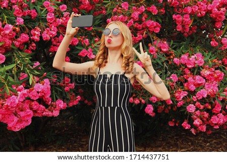 Beautiful young woman taking selfie picture by smartphone blowing red lips sending sweet air kiss over flowers roses background