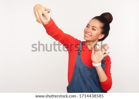 Hello! Portrait of friendly stylish girl with hair bun in denim overalls waving hi gesture and making video call on mobile phone, using gadget for online communication. studio shot, white background