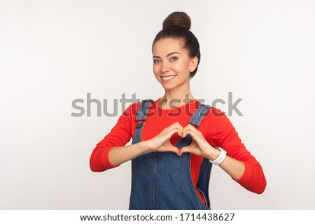 I love you! Portrait of attractive joyful girl with hair bun in denim overalls making heart shape with fingers, showing gesture of affection, care and generosity. indoor studio shot, white background