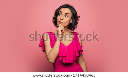 Hard choice. Close-up photo of a gorgeous brunette lady in a pink dress, who is looking to the right and touching her chin with her right hand, thinking over something.