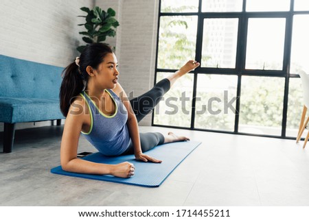 Strong fitness girl in athletic exercise clothes doing a side kick workout while lying down. Asian woman training at home in her living room with cozy sofa home interior setting with copy space Royalty-Free Stock Photo #1714455211