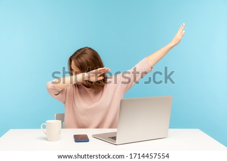 Dabbing trends. Overjoyed woman showing dab dance gesture, performing internet meme of success, sitting at workplace with laptop, home office job. indoor studio shot isolated on blue background Royalty-Free Stock Photo #1714457554