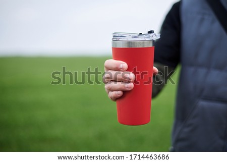 Close-up picture of male hand, holding red travel mug with hot tea coffee drink outside in the field in spring summer. Healthy lifestyle concept. Royalty-Free Stock Photo #1714463686
