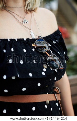 Close-up picture of woman's body, wearing black dotted dress, silver chains, brown leather belt and sunglasses. Summer outfit details.