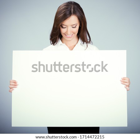 Smiling looking down woman showing blank sign board with empty copy space for some slogan, text. Business ad concept. Multiracial Asian Chinese / Caucasian model over grey background.