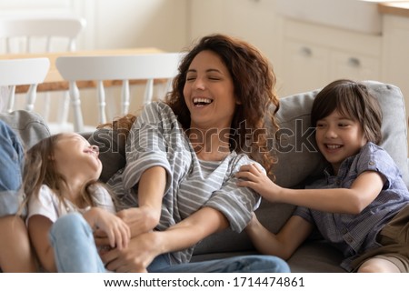Excited young nanny babysitter tickling small children boy girl, having fun on sofa in modern studio living room. Happy small kids brother sister laughing, enjoying daycare time with pleasant mother. Royalty-Free Stock Photo #1714474861