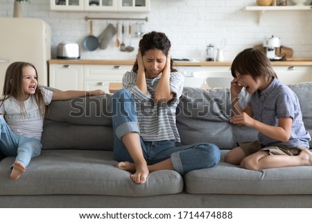Desperate mother nanny babysitter annoyed by hyperactive loud screaming children, suffering from strong headache. Stressed young mommy irritated by bad kids siblings behavior, covering ears. Royalty-Free Stock Photo #1714474888
