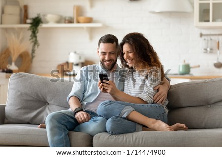 Happy young wife showing funny video in social network on cellphone to laughing husband. Smiling spouses shopping online web surfing, relaxing together on sofa in modern studio kitchen living room. Royalty-Free Stock Photo #1714474900