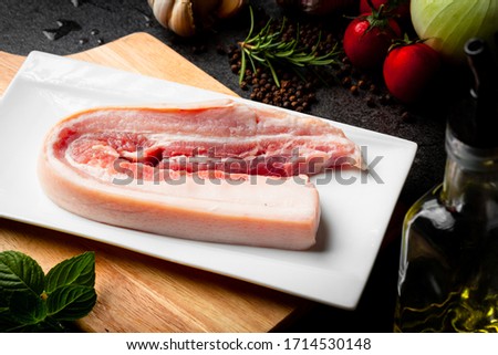 raw pork belly with garlics, tomatoes, rosemarry,Olive oil, pepper, bbq sauce and on stone black background, dark picture style.