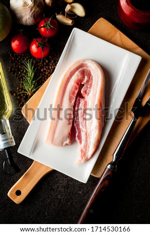 raw pork belly with garlics, tomatoes, rosemarry,Olive oil, pepper, bbq sauce and carving fork on stone black background, dark picture style.