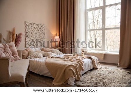 Bedroom in beige tones with a large bed and a large window through which a lot of light penetrates. Minimal style in the interior. Convenient and practical location of furniture, cozy view.
