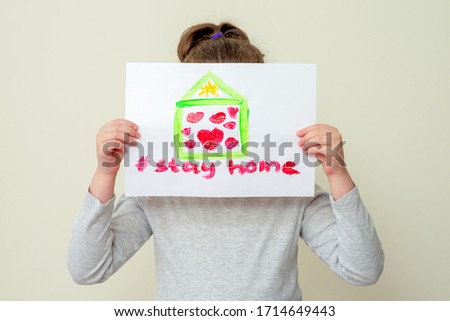 Little girl covers his face a picture of a house with hearts and words Stay Home on sheet of paper, close-up. Stay home concept.