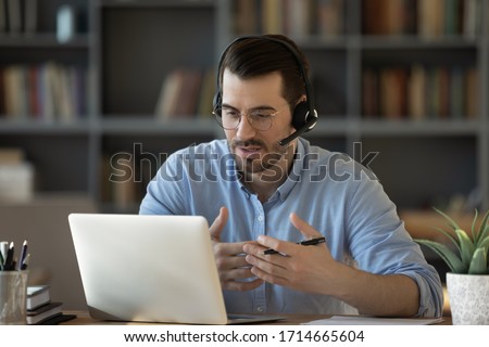 Confident man teacher coach wearing headset speaking, holding online lesson, focused student wearing glasses looking at laptop screen, studying, watching webinar training, listening to lecture Royalty-Free Stock Photo #1714665604