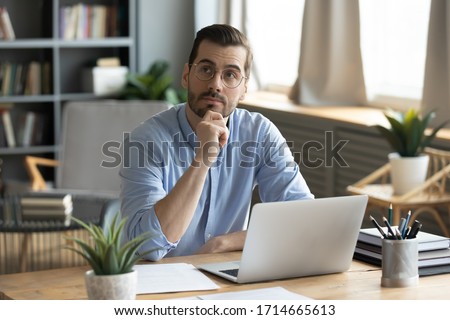 Thoughtful businessman wearing glasses touching chin, pondering ideas or strategy, sitting at wooden work desk with laptop, freelancer working on online project, student preparing for exam at home Royalty-Free Stock Photo #1714665613