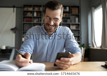 Smiling businessman wearing glasses using phone, writing important information in notebook, sitting at desk, happy man holding smartphone, making notes in personal daily planner, planning work day Royalty-Free Stock Photo #1714665676