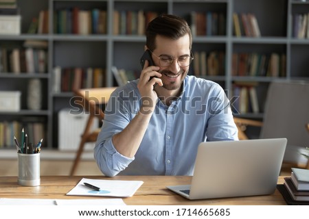 Smiling businessman wearing glasses talking on phone, sitting at desk with laptop, friendly manager consulting customer by phone, happy man chatting with friends distracted from work