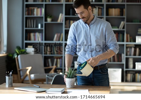 Confident businessman wearing glasses watering plant on work desk, serious focused man holding plastic pot with water, standing near table in modern office cabinet, new startup project