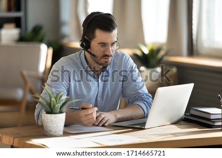 Focused man wearing headset writing notes, serious young student wearing glasses studying, looking at screen, watching webinar training, listening to lecture, mentor coach teaching online