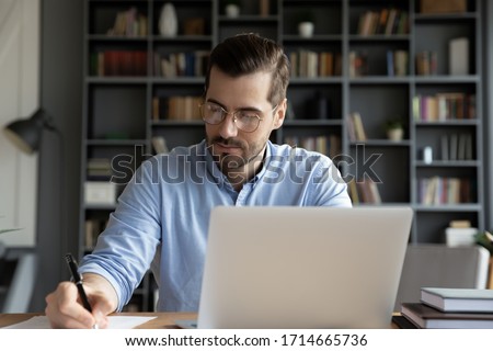 Confident businessman wearing glasses writing notes or financial report, sitting at desk with laptop, focused serious man working with paper documents, student studying online, research work Royalty-Free Stock Photo #1714665736