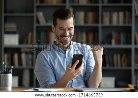 Excited smiling man wearing glasses holding phone, reading good news in message, happy young male looking at screen, celebrating online lottery win, showing yes gesture, sitting at work desk Royalty-Free Stock Photo #1714665739