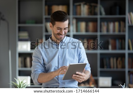 Smiling businessman wearing glasses using computer tablet, standing in modern cabinet, happy positive man enjoying leisure time, playing game, chatting in social network, browsing apps Royalty-Free Stock Photo #1714666144