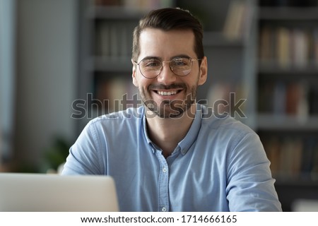 Head shot portrait smiling businessman student worker wearing glasses looking at camera, happy satisfied young man sitting at work desk with laptop, posing for photo in modern cabinet