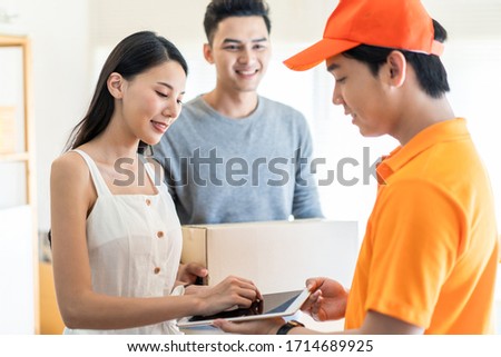 Asian postman or deliveryman get box package from customer home and send to destination. Woman client signs delivery form on tablet. Social distancing work during Covid outbreak. Door to door service.