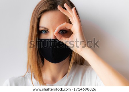 Beautiful close-up portrait of a young woman in a black medical mask and white T-shirt on white background, makes okay sign with hand. Quarantine, stop coronavirus pandemic, covid-19. Stay home
