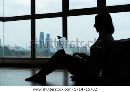 Siluet girl working on a laptop and drinking coffee, sitting on the floor near the bed by the panoramic window with a beautiful view from the high floor. Stylish modern interior. A cozy workplace