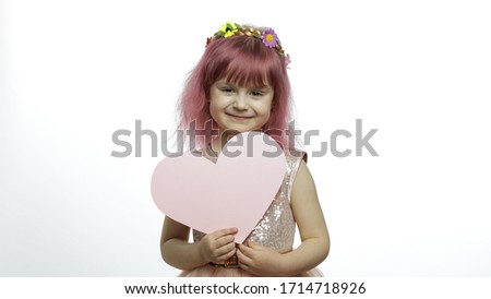 Smiling little girl princess holds a cut pink paper heart in her hands. White background. Cute child enjoying happy childhood, showing love sign. Mothers day, Valentines day concept