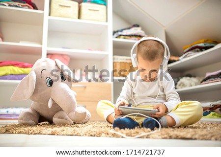 Adorable little boy with headphones sitting on the floor with favorite plush toy and watching cartoons on tablet