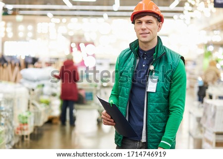 expert in his field warehouse worker stand in the market look at camera and smile, wearing green uniform and protective helmet