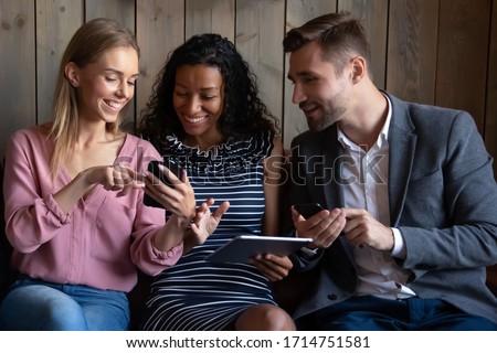 Happy multiracial young people sit on couch have fun using electronic gadgets together, smiling diverse multiethnic friends relax in cafe laugh joke showing pictures on devices, technology concept