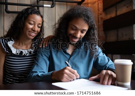 Excited couple sit in coffee shop sign paper document make agreement, happy African American  man and woman close deal with wedding planner or consultant in cafe