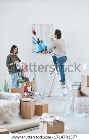 Young man in casualwear standing on stepladder by wall and hanging abstract painting while his wife looking at picture in frame