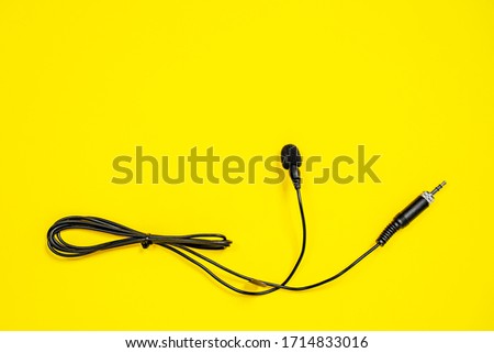Lapel microphone with a bundled cord on yellow background, top view. Essential vlogging and journalism tools. Piece of audio equipment designed to record sounds and speech