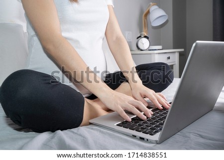 side view of pregnant woman in white t-shirt and black pants sitting on bed while using laptop