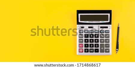 Large silver calculator with gray and black buttons and black pen on a yellow background. Conceptual photo of calculations, accounting, computing, profit, loss, tax with place for text. Royalty-Free Stock Photo #1714868617